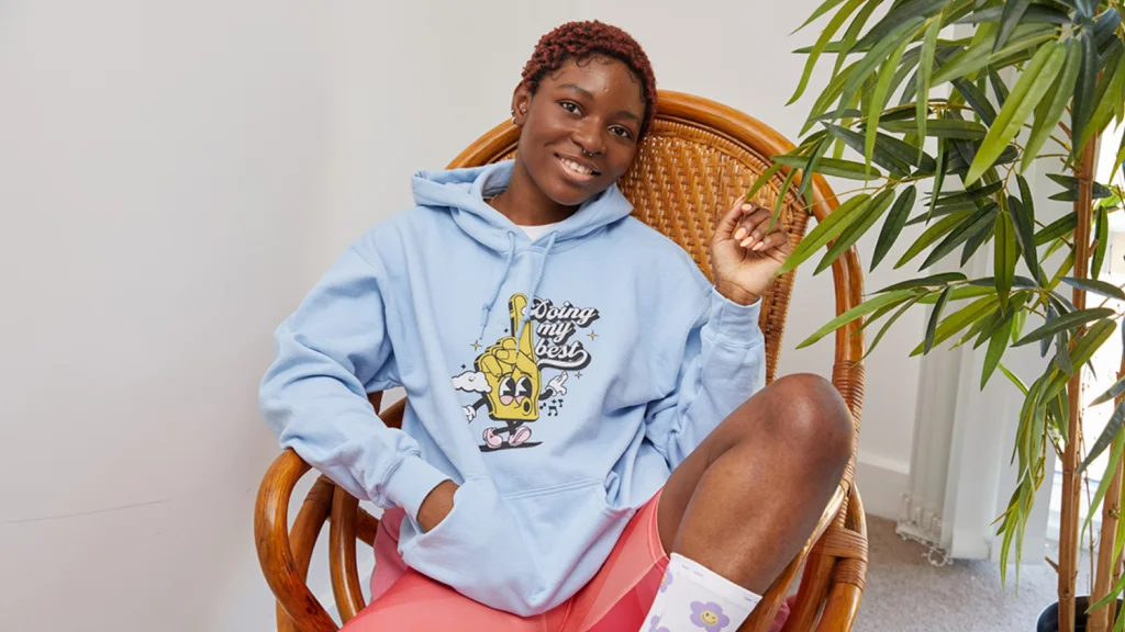 Gildan hoodie vs sweatshirt A quick comparison A smiling woman with short hair sitting in a wicker chair, wearing a light blue hoodie with a playful cartoon design and pink shorts.