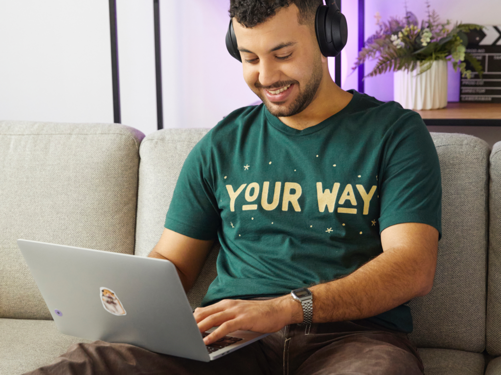 A man with headphones smiles as he types on a laptop, seated on a gray sofa. He wears a green 'Your way” t-shirt.
