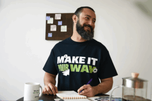 A bearded man smiles while making notes, seated at a table. He wears a "Make It Your Way" t-shirt.
