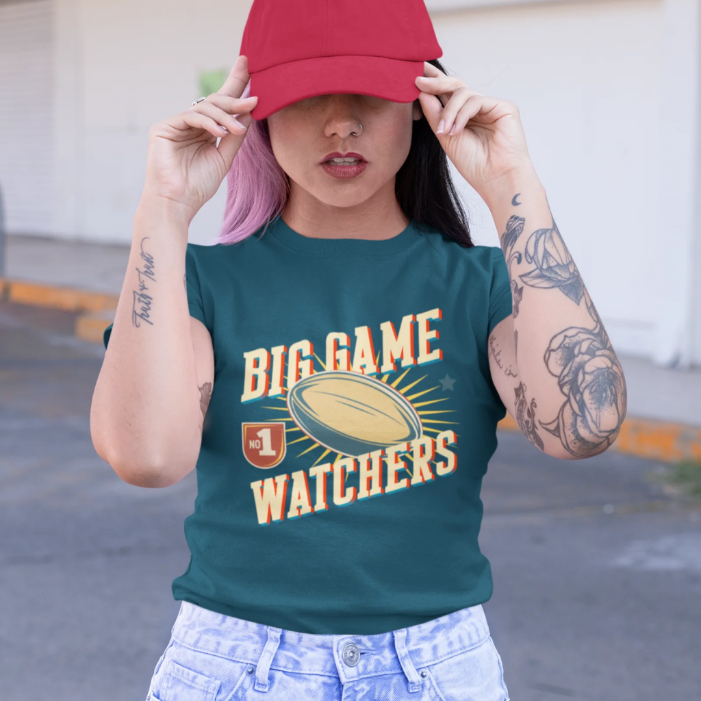 A woman with tattoos adjusts a red cap, wearing a teal shirt saying "Big Game Watchers" with a football graphic.