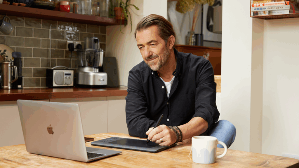 A man in a black shirt sits at a wooden kitchen table, using a tablet with a stylus. An open laptop and mug sit nearby.