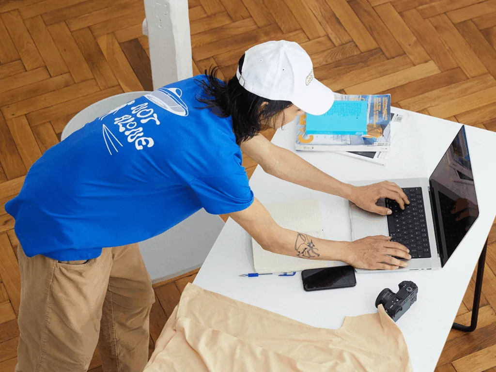 A man in a blue shirt and white cap leans over a desk, typing on a laptop. A notebook, phone, camera, and papers are nearby.
