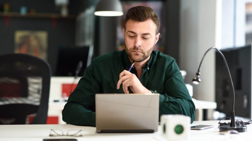 A man in a dark green shirt sitting in front of a laptop and thinking.