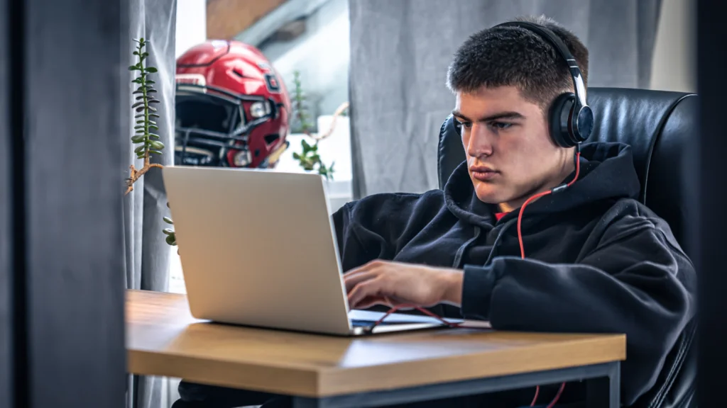 A focused young man in headphones works on a laptop at a desk. A red football helmet is in the background.