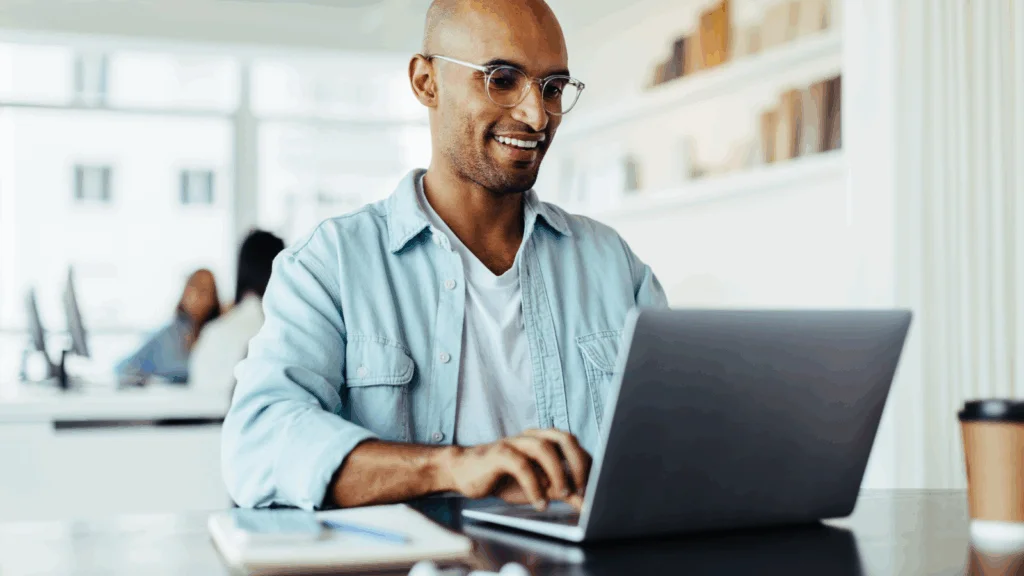 A smiling man wearing glasses works on a laptop, searching for how to start a print-on-demand business.