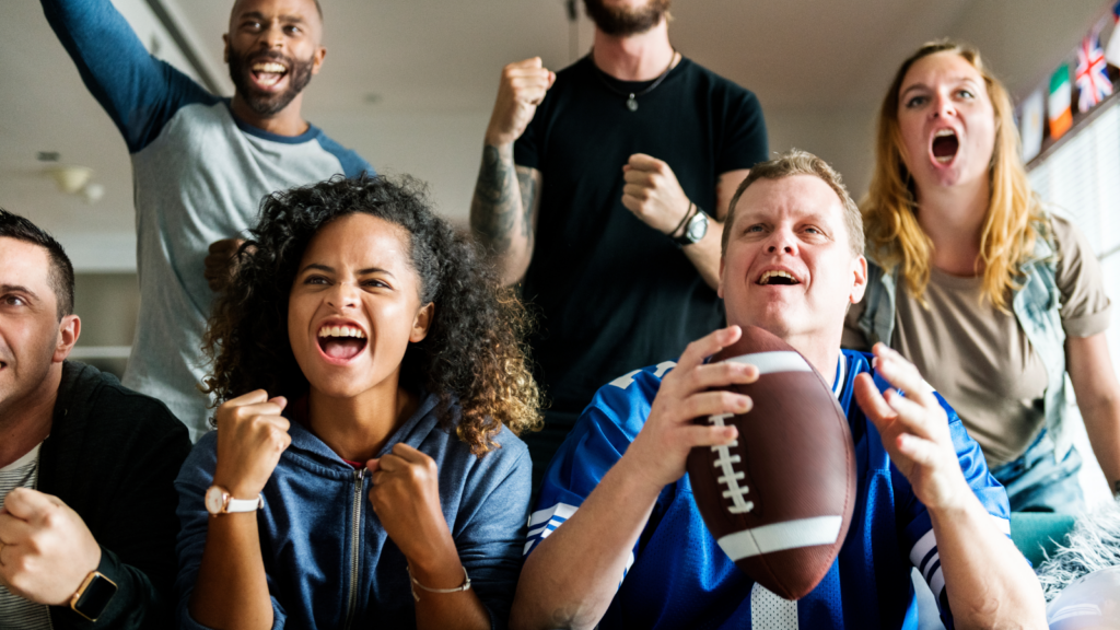 A diverse group of six people excitedly watches a game on TV, cheering with fists raised. One holds a football.