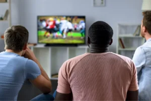Three men sit on a couch watching a football game on TV, with a blurred action scene on screen.