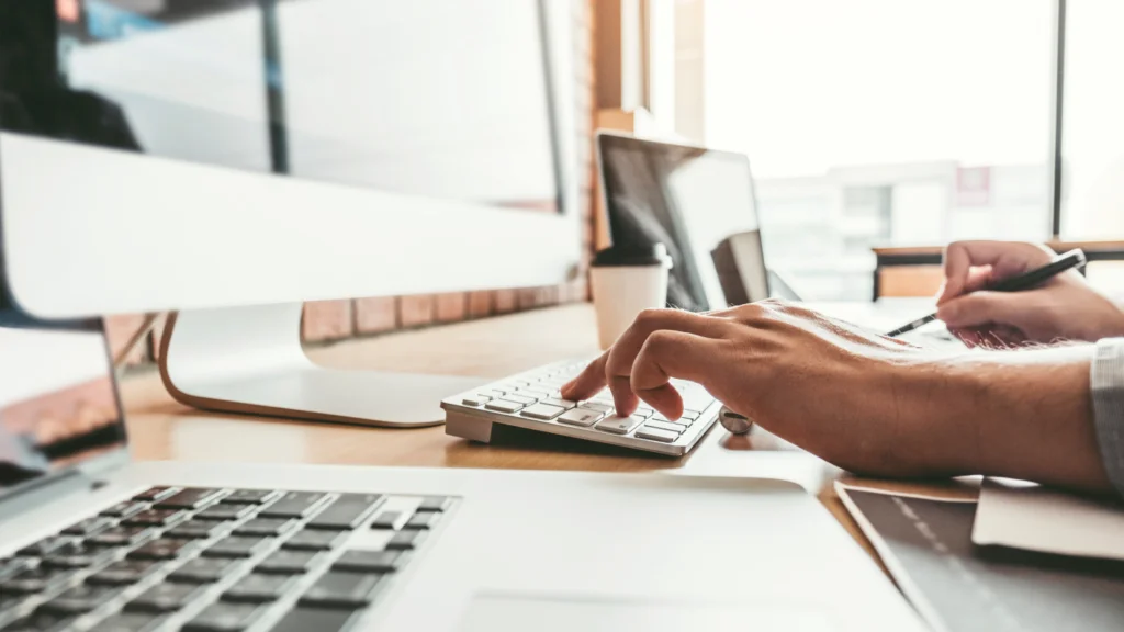 A man typing on a keyboard, “how to design a t-shirt”, beside a modern desk setup with multiple monitors and a laptop.