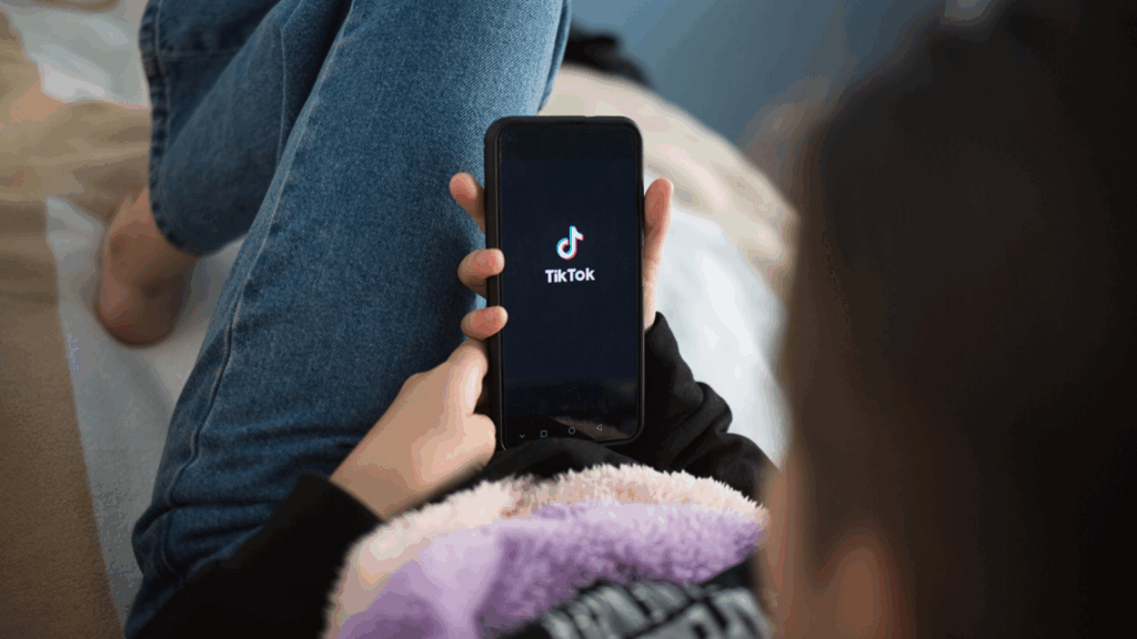 A person sitting on a bed, holding a cell phone displaying the TikTok logo.
