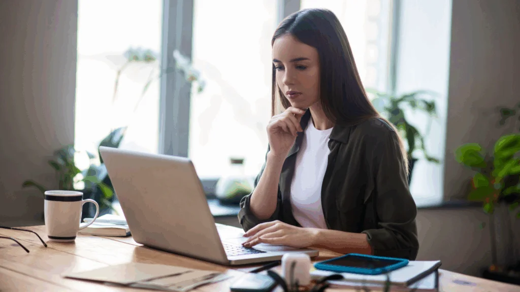 A woman seated at a desk, focused on her laptop, with a notepad and pen beside her.