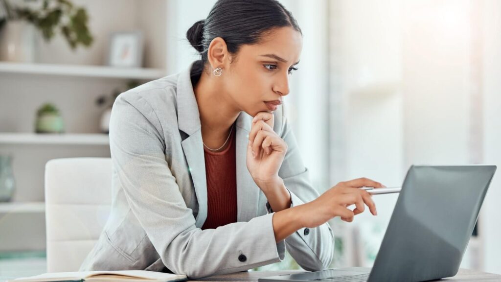 A woman in a gray blazer looking at a laptop screen and pointing with a pen.