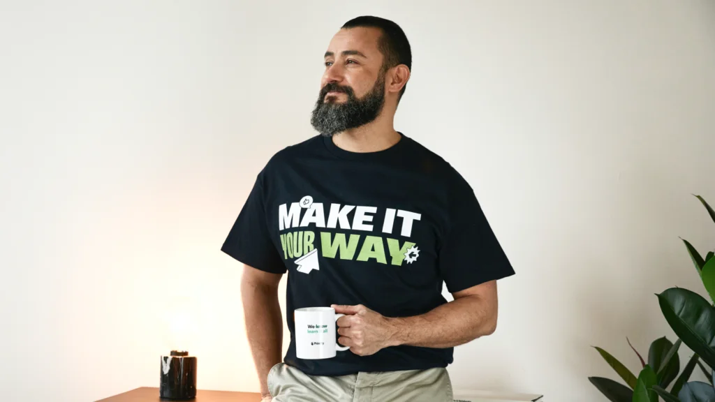A man with a beard wearing a black "Make It Your Way" t-shirt and holding a mug, stands confidently near a plant and lamp.