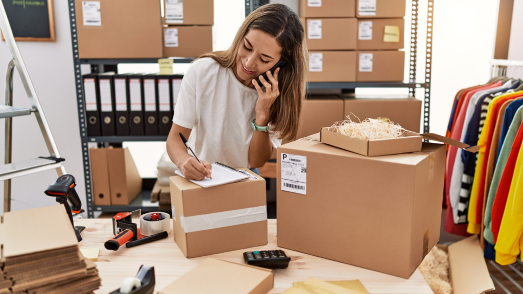 Choose production and shipping methods A woman sits at a desk, focused on her computer, surrounded by boxes and papers, engaged in her work.
