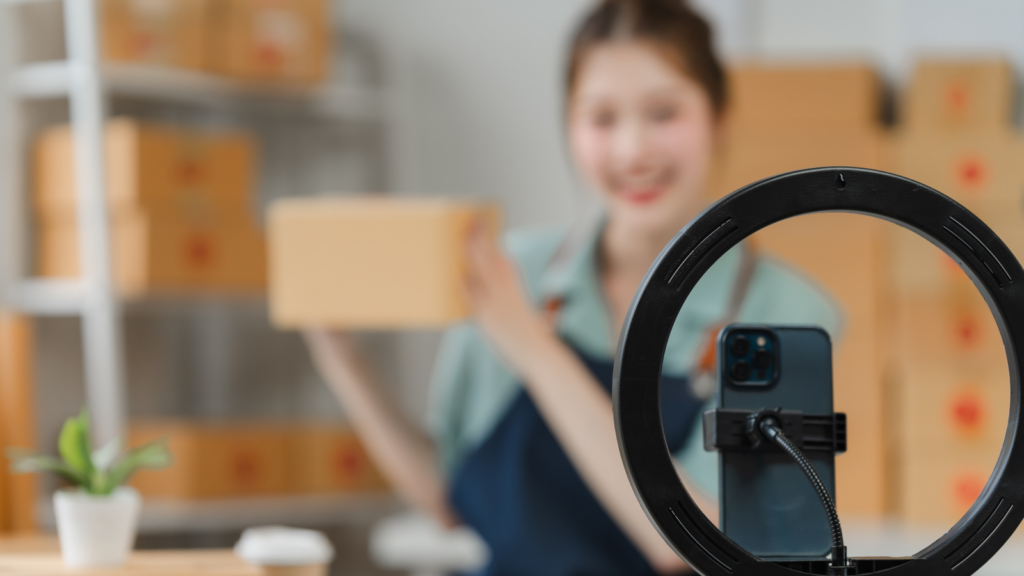 Collaborate with influencers A mobile phone on a stand in front of a blurred woman holding a box and filming herself.