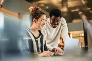 Two professionals engaged in a discussion while working on a laptop in an office environment.