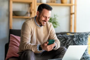 A smiling man sitting on a couch with a laptop in front of him and a mobile phone in his hands.
