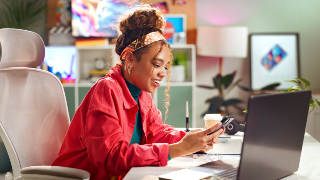 A woman holding a mobile phone in her hand and smiling while sitting in front of a laptop.