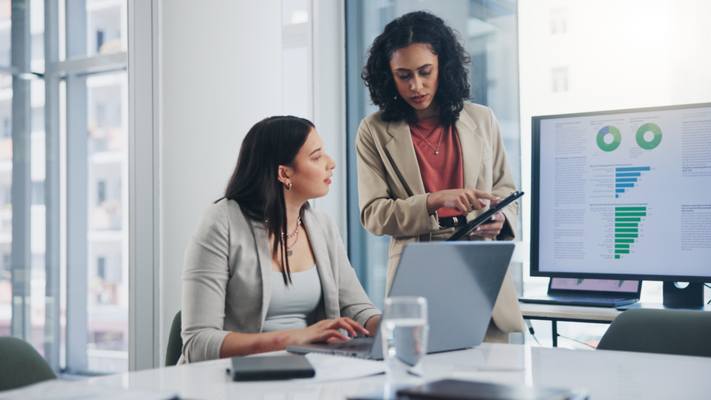Optimize your Shopify store In an office, two women engage in discussion as they review information on a laptop.