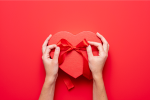 Hands gently holding a red heart-shaped box against a vibrant red background, symbolizing love and affection.