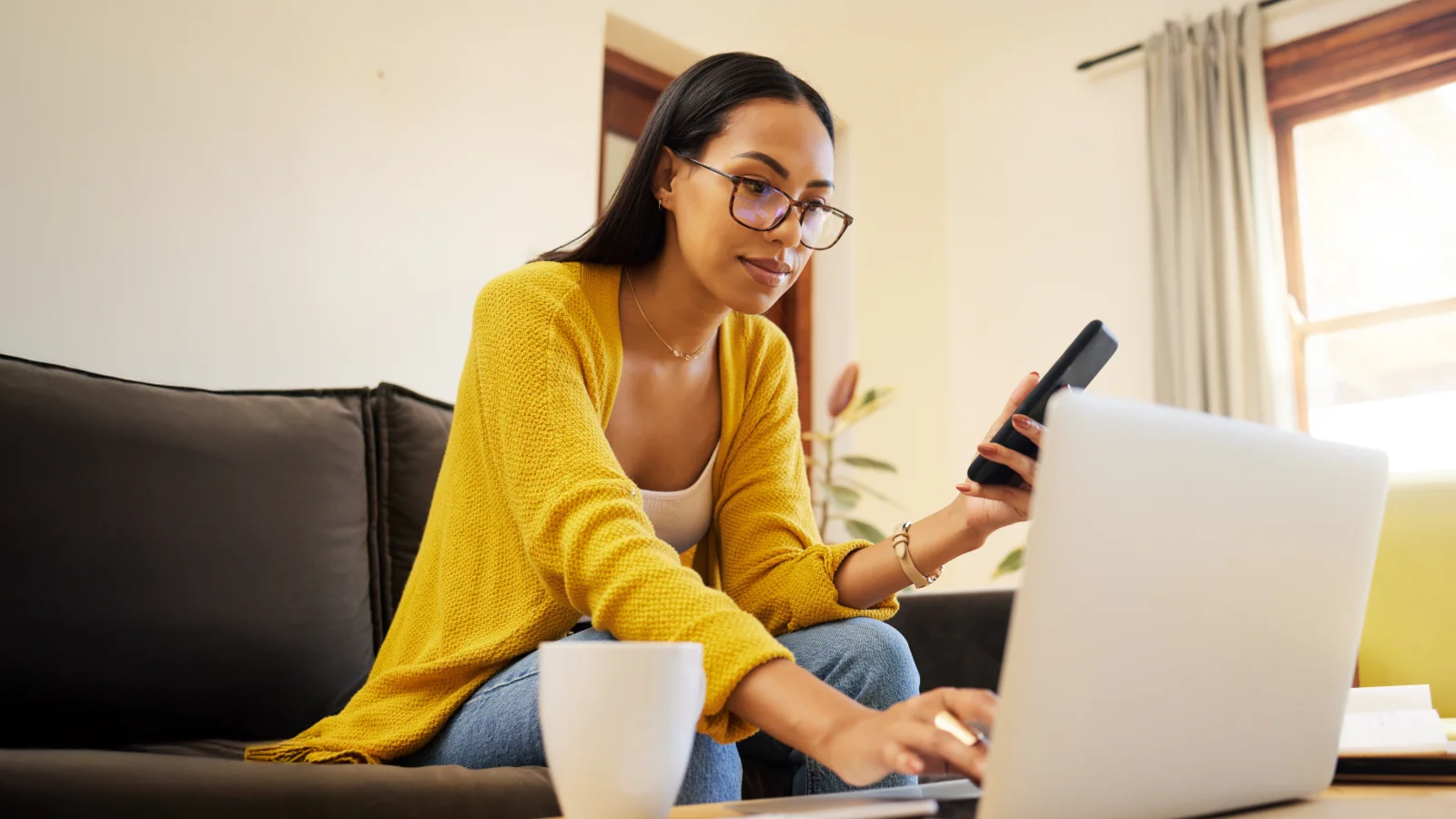 A woman wearing glasses sits on a couch, working on a laptop.