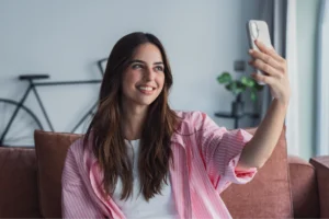 A smiling woman taking a selfie while sitting on a couch. She's wearing a pink shirt, with a bicycle and a plant in the background.