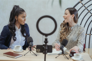Two women sitting in front of a mobile phone, recording a podcast.