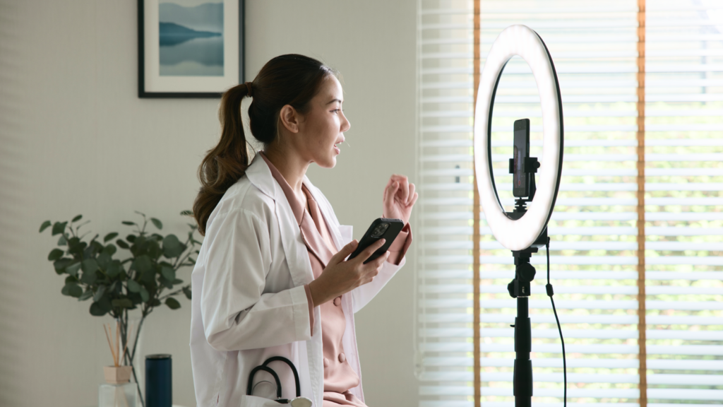 A woman in a white coat films a video using a smartphone mounted on a ring light. She gestures while speaking.