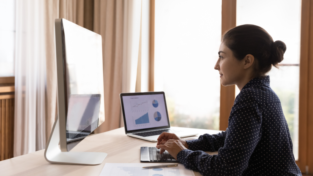 A woman sitting at a desk and looking at a monitor with a laptop beside her.