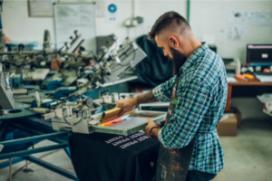 A man diligently working on a t-shirt in a factory setting, focused on his task amidst the production environment.