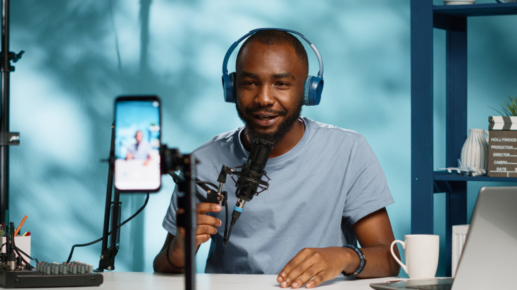 A man in blue headphones speaks into a microphone while recording himself with a smartphone.