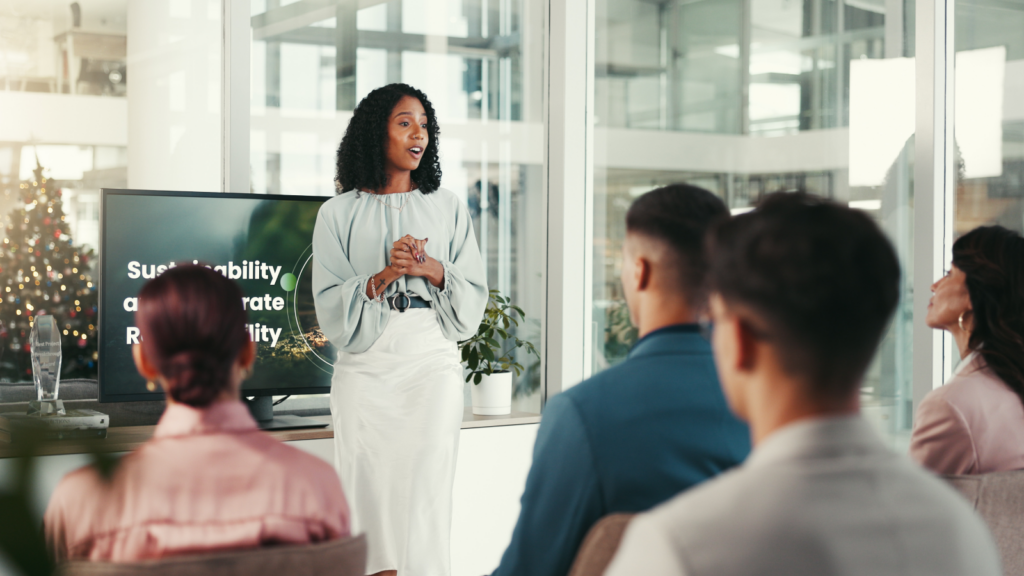 Online workshops or coaching A woman stands confidently at the front, presenting to an engaged audience of diverse individuals seated in a conference room.