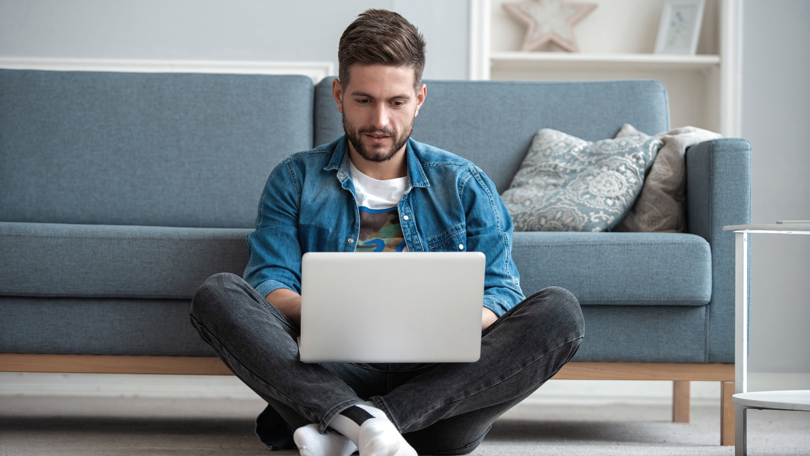 Research the sticker market and identify your target audience A man sitting on the floor in front of a couch and using a laptop that he has on his lap.