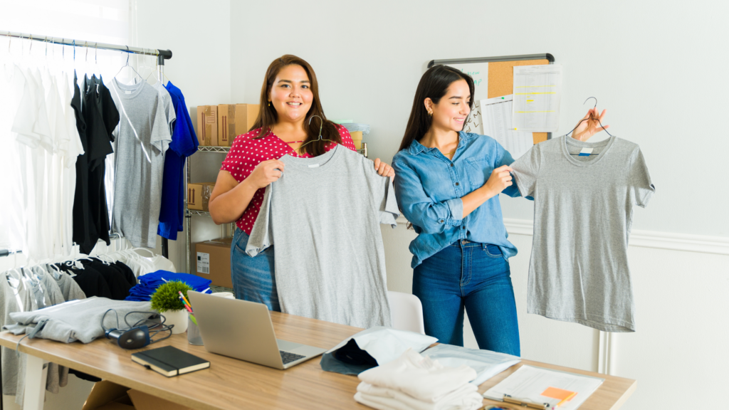 Start a print on demand business Two women are standing in a room, each holding a shirt and learning how to make money as stay-at-home moms.