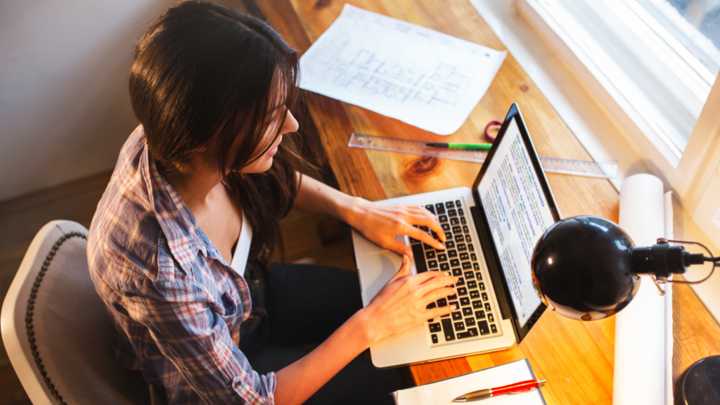 A woman using a laptop with different plans and papers surrounding the laptop.