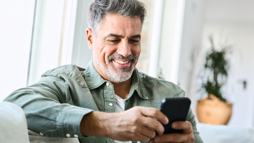 A smiling man with gray hair and beard wearing a green shirt, sitting indoors and looking at his smartphone. A plant is blurred in the background.
