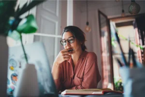 A woman wearing glasses is seated at a desk, focused on her laptop while working diligently.