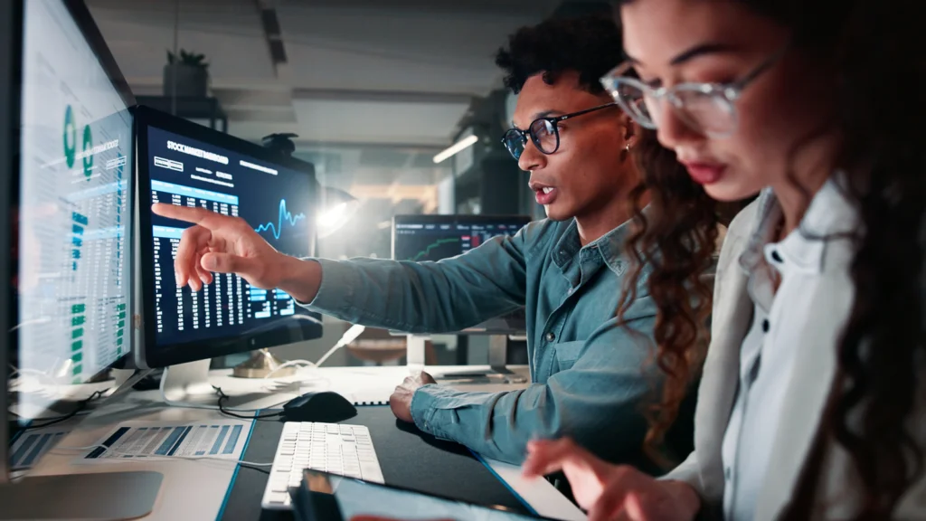 Two people work in a dimly lit office, focused on financial data displayed on multiple computer screens.