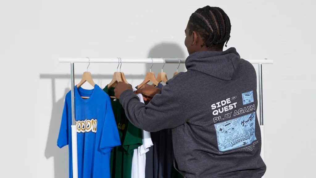 A man in a graphic hoodie looks at colorful t-shirts on a clothing rack.