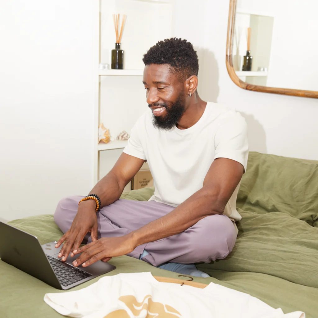A man with a beard smiles while using a laptop to learn how to sell t-shirts on Etsy, sitting cross-legged on a bed.