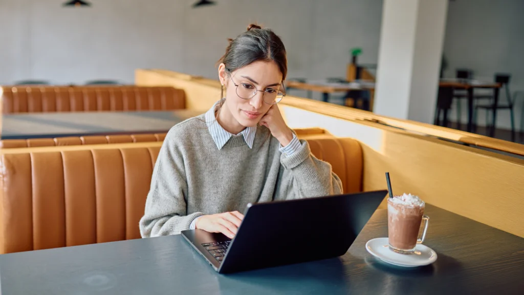 A woman with glasses sits in a cozy café, focused on her laptop. She wears a gray sweater with a drink beside her.