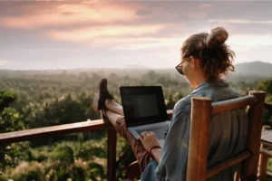 A woman sitting on a balcony with beautiful scenery, writing a blog about how to make money while traveling.