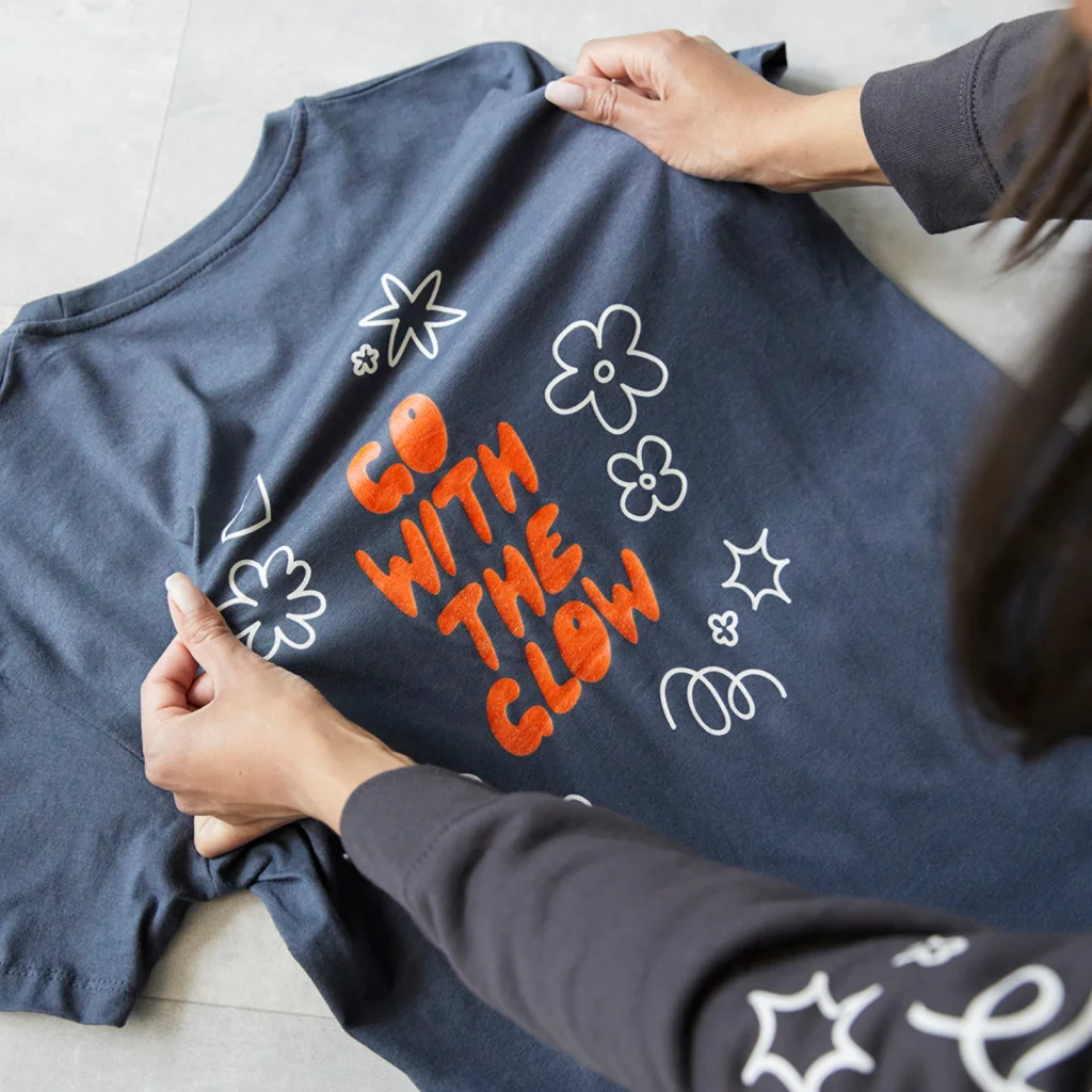 Hands hold a dark blue t-shirt featuring the phrase "GO WITH THE GLOW" in bold orange letters, surrounded by white floral designs.