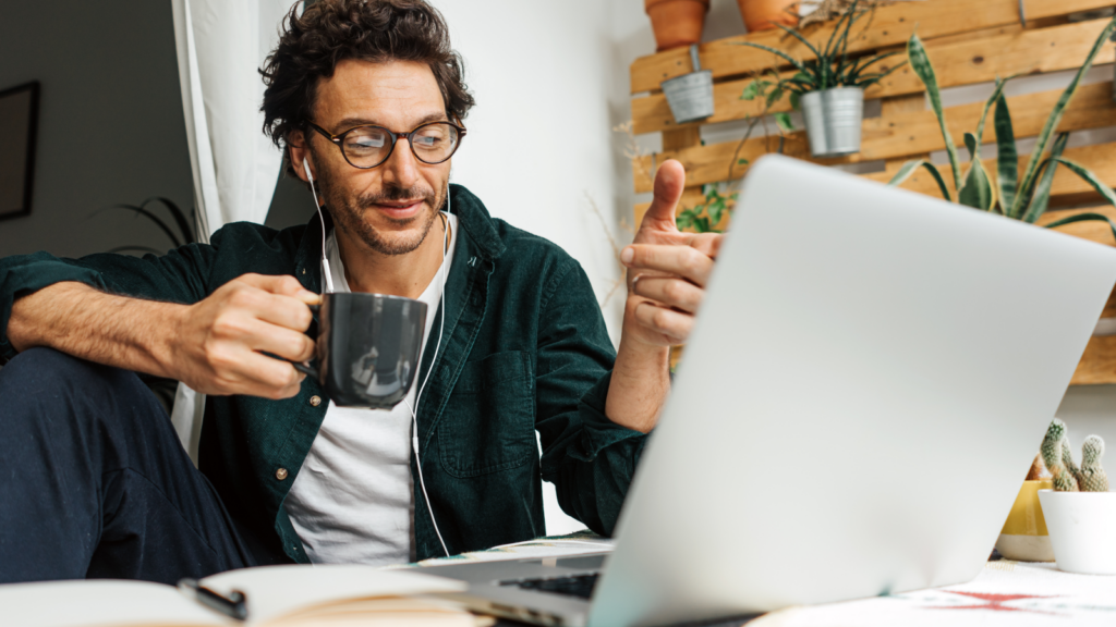 A man with glasses and earphones gives a thumbs-up while holding a mug, smiling at a laptop. Plants and shelves in the background.