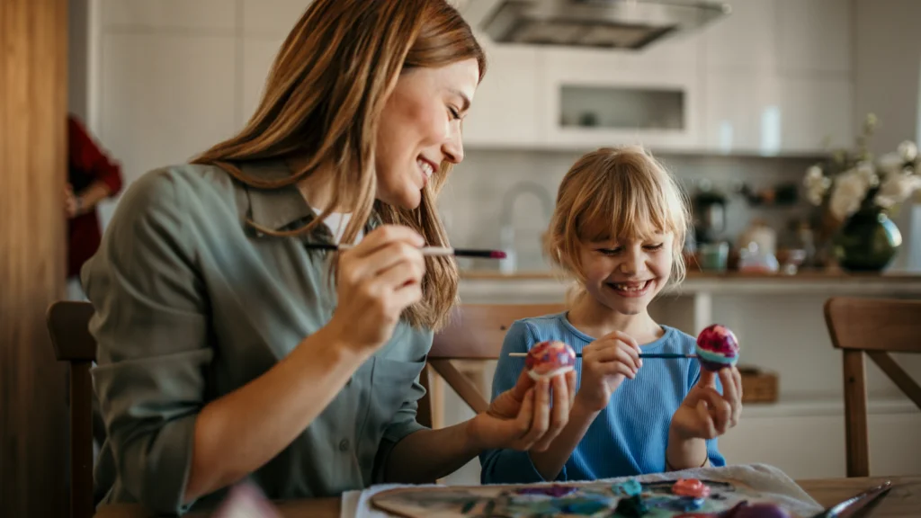 A mother and daughter smile while painting eggs at a kitchen table, conveying joy and creativity.