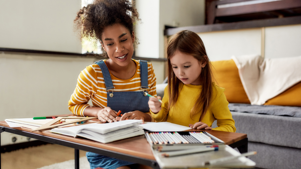 A woman babysitting a young girl sits at a table drawing with colored pencils.
