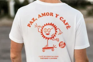 A man in a t-shirt displaying the words "paz amor y cafe," smiling and standing against a neutral background.