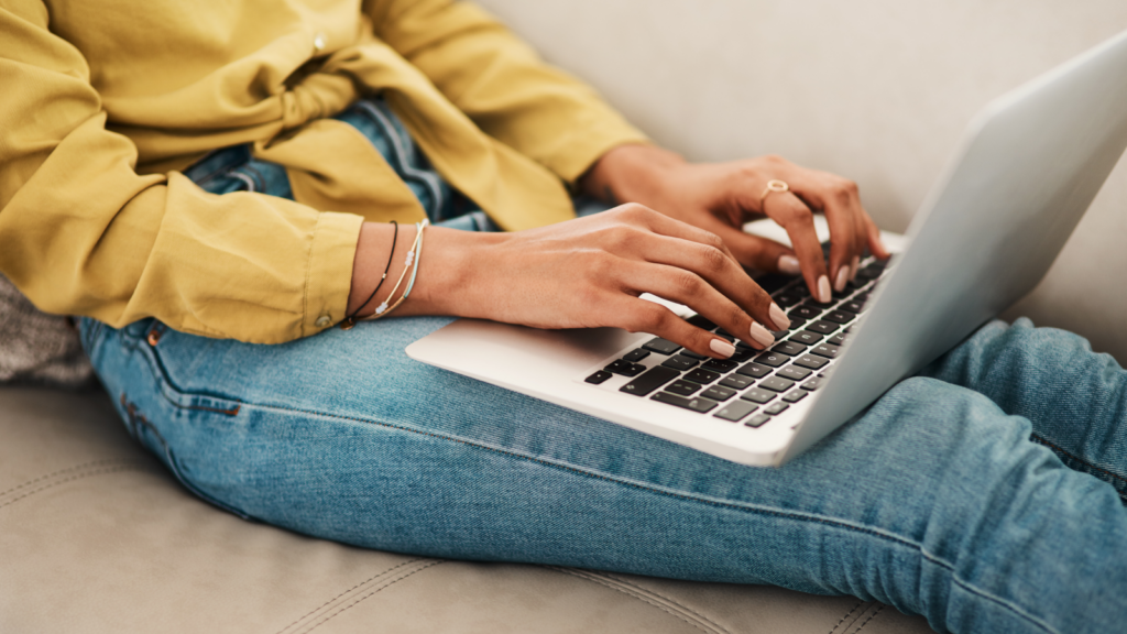 A woman in a yellow blouse and blue jeans, sitting on a couch with a laptop on her lap, typing.