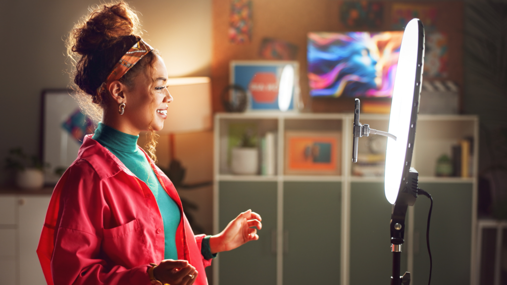 A woman in a vibrant setting speaks enthusiastically in front of a ring light. She wears a bright pink jacket and a colorful headband.