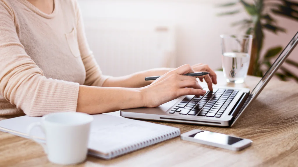 A person types on a laptop at a wooden desk, surrounded by a notebook, coffee mug, smartphone, and glass of water.