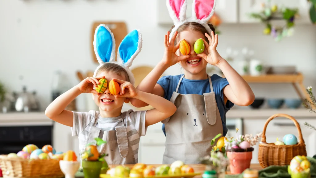 Two children in aprons wear bunny ears, holding colorful eggs to their faces in a kitchen. Easter decorations and baskets fill the scene.